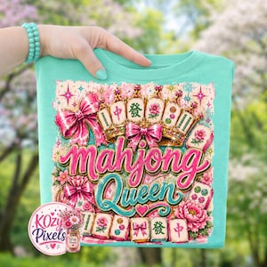 May include: A turquoise t-shirt featuring a colorful graphic design. The design includes the words "Mahjong Queen" in pink and teal script, surrounded by pink bows, floral elements, and Mahjong tiles. The design also includes gold crowns and Chinese characters.