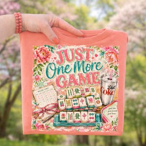 May include: Peach-colored sweatshirt featuring a colorful illustration of a Mahjong game, a Diet Coke, and the text "Just One More Game." The design includes floral accents and a vintage aesthetic, perfect for game lovers.