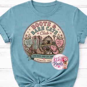 May include: A dusty blue t-shirt featuring a circular graphic with the text Boots & Barn Cats Club. The design includes a pair of cowboy boots, a barn, a cat, a pink cowboy hat, and two hearts with text. A small circular logo for Kozy Pixels is present.