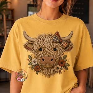 May include: A mustard yellow t-shirt featuring a graphic of a Highland cow with a textured, yarn-like appearance. The cow has a plaid bow on one horn and is surrounded by small flowers. The text Kozy Pixels by Sheila is printed in the corner.