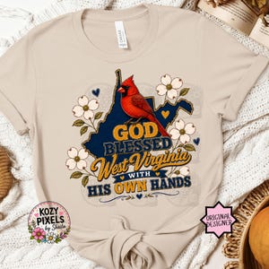 May include: A beige t-shirt featuring a graphic of a red cardinal bird perched on a dark blue map of West Virginia. The design includes white dogwood flowers and the text "GOD BLESSED West Virginia WITH HIS OWN HANDS" in yellow and blue lettering.