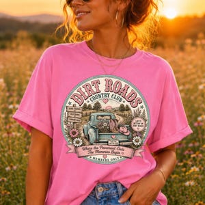 May include: A bright pink short-sleeve t-shirt featuring a circular graphic with a vintage truck, cowboy boots, and flowers. The text reads Dirt Roads Country Club, Life's Better On Dirt Roads, Where The Pavement Ends The Memories Begin, Members Only.