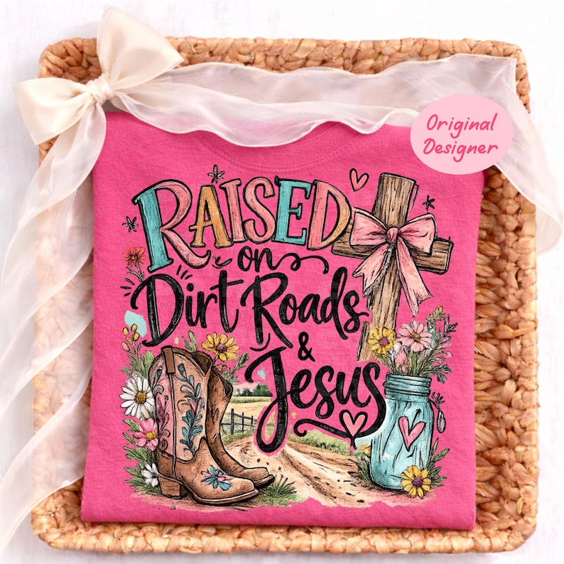 K&ouml;nnte beinhalten: Ein leuchtend pinkes T-Shirt mit einem Grafikdruck und dem Text Raised on Dirt Roads & Jesus. Das Design zeigt ein Holzkreuz mit einer rosa Schleife, ein Paar braune Cowboystiefel, ein blaues Einmachglas mit Blumen und bunte Wildblumen.