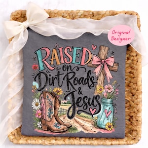 K&ouml;nnte beinhalten: Ein dunkelgraues T-Shirt mit einem Grafikdruck und dem Text "Raised on Dirt Roads & Jesus". Das Design zeigt ein Holzkreuz mit einer rosa Schleife, ein Paar braune Cowboystiefel, ein blaues Einmachglas mit Blumen und bunte florale Akzente.