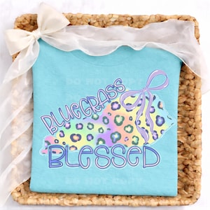 Peut inclure: Un t-shirt bleu clair avec l'inscription "Bluegrass Blessed" en violet et bleu. Le motif comprend un imprimé léopard aux couleurs pastel et un nœud violet. Le t-shirt est présenté dans un panier en osier avec un ruban blanc.