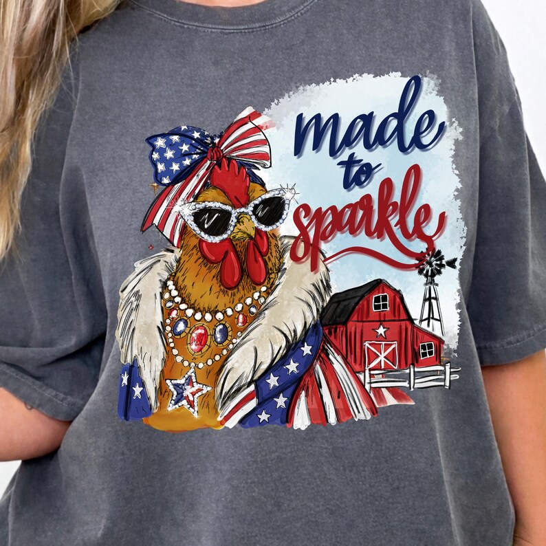 Op de afbeelding: Donkergrijs t-shirt met een cartoonkip met een zonnebril, een patriottische strik en sieraden. De afbeelding bevat de tekst "made to sparkle" met een rode schuur en windmolen op de achtergrond. Het ontwerp bevat rode, witte en blauwe elementen.