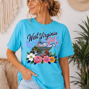 Może przedstawiać: Jasnoturkusowy t-shirt z krótkim rękawem z nadrukiem graficznym i napisem West Virginia Girl. Wzór przedstawia zarys mapy Wirginii Zachodniej, most, wodospad, czerwonego kardynała oraz kolorowe akcenty kwiatowe.