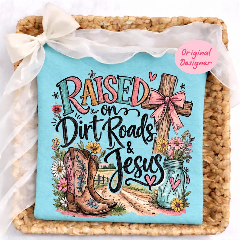 K&ouml;nnte beinhalten: Ein hellblaues T-Shirt mit einem Grafikdruck und dem Text "RAISED on Dirt Roads & Jesus". Das Design zeigt einen braunen Cowboystiefel, ein Holzkreuz mit einer rosa Schleife, ein Einmachglas mit Blumen und eine Landschaft mit einem Feldweg.