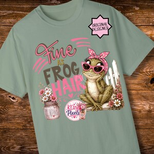 K&ouml;nnte beinhalten: Hellgr&uuml;nes T-Shirt mit einer skurrilen Frosch-Grafik, die eine Sonnenbrille und eine rosa Schleife tr&auml;gt. Der Text lautet "Fine as Frog Hair" in Rosa und Grau. Ein Glas mit Blumen und das Logo "Kozy Pixels" sind ebenfalls abgebildet.
