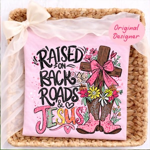 Puede incluir: Una camiseta rosa con el texto "RAISED ON BACK ROADS & JESUS" en una mezcla de fuentes y colores. El diseño incluye una cruz de madera con un lazo rosa, flores y botas de vaquero rosas. La camiseta se presenta en una cesta tejida con una cinta blanca.