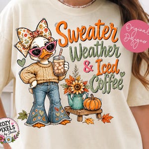 May include: A cream-colored t-shirt featuring a graphic of a duck wearing a floral bow, pink sunglasses, and a tan sweater with blue jeans. The text reads Sweater Weather & Iced Coffee, accompanied by a sunflower bouquet and a pumpkin.