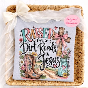K&ouml;nnte beinhalten: Ein hellgraues T-Shirt mit einer bunten Grafik und dem Text "Raised on Dirt Roads & Jesus". Das Design zeigt ein Holzkreuz mit einer rosa Schleife, ein Paar braune Cowboystiefel, ein blaues Einmachglas mit Blumen und einen Feldweg.