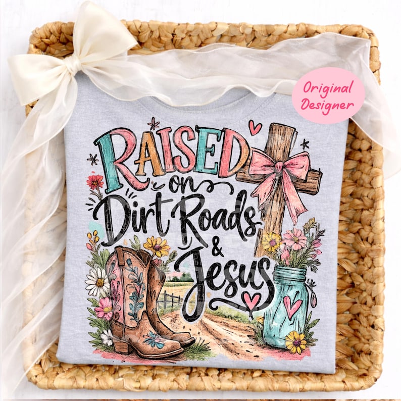 K&ouml;nnte beinhalten: Ein hellgraues T-Shirt mit einer bunten Grafik und dem Text "Raised on Dirt Roads & Jesus". Das Design zeigt ein Holzkreuz mit einer rosa Schleife, ein Paar braune Cowboystiefel, ein blaues Einmachglas mit Blumen und einen Feldweg.