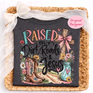 K&ouml;nnte beinhalten: Ein dunkelgraues T-Shirt mit einem Grafikdruck und dem Text Raised on Dirt Roads & Jesus. Das Design zeigt einen braunen Cowboystiefel, ein Holzkreuz mit einer rosa Schleife, ein blaues Einmachglas mit Blumen und eine Feldweg-Landschaft.