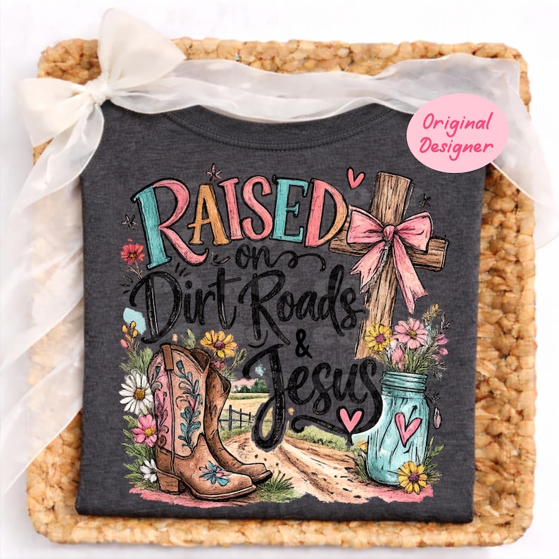 K&ouml;nnte beinhalten: Ein dunkelgraues T-Shirt mit einem Grafikdruck und dem Text Raised on Dirt Roads & Jesus. Das Design zeigt einen braunen Cowboystiefel, ein Holzkreuz mit einer rosa Schleife, ein blaues Einmachglas mit Blumen und eine Feldweg-Landschaft.
