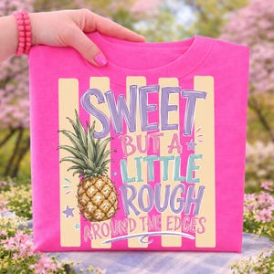 May include: A bright pink t-shirt with a pineapple graphic and the text "SWEET BUT A LITTLE ROUGH AROUND THE EDGES". The pineapple is detailed with brown and green colors. The text is in purple, blue, and green.