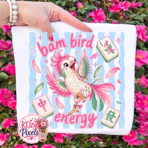 May include: White square fabric with a colorful illustration of a parrot with pink, green, and white feathers. The text "bam bird energy" is above and below the parrot. Mahjong tiles and feathers are also featured in the design.
