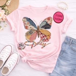 May include: A light pink t-shirt featuring a large butterfly graphic composed of various patchwork fabric patterns in shades of blue, red, yellow, and white. The word Dolly is written below the butterfly in a script font. A pink circle reads Original Designer.