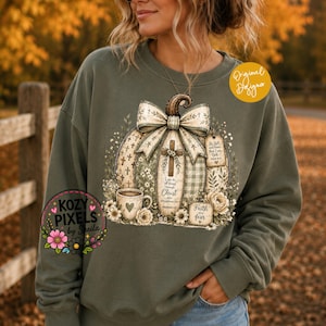 May include: An olive green crewneck sweatshirt featuring a large graphic of a pumpkin decorated with a bow, a cross, and floral accents. The design includes text that reads "I can do all things through Christ who strengthens me," "Faith over fear," and "Be still and know that I am God."