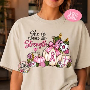 May include: A beige t-shirt featuring a graphic print of two pumpkins, one white and one pink, decorated with a pink ribbon, flowers, and a small sign with a cross. The text reads She is CLOTHED WITH Strength and PROVERBS 31:25.