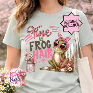 Könnte beinhalten: Hellgrünes T-Shirt mit dem Schriftzug "Fine as Frog Hair" in Rosa und Grau. Das Design zeigt einen Cartoon-Frosch mit Sonnenbrille und rosa Kopftuch, der sich auf einem Holzzaun abstützt. Ein Glas mit Blumen ist ebenfalls abgebildet.