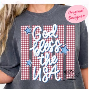 以下が含まれることがあります： 「God bless the USA」の文字が青と白の筆記体で書かれた濃いグレーのTシャツ。デザインには青い星のアクセントと赤と白のチェック柄が含まれています。「Original Designer」の文字がピンクの円で囲まれています。