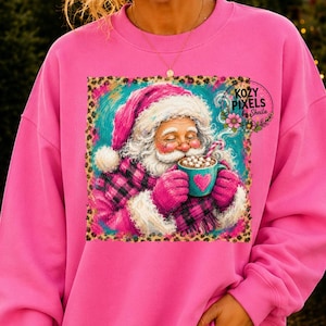 May include: A bright pink sweatshirt featuring a square graphic of Santa Claus wearing a pink and black plaid coat and hat, holding a teal mug with a pink heart. The graphic includes the text Kozy Pixels by Sheila and a floral border.
