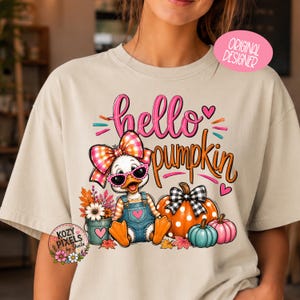 Puede incluir: Una camiseta beige con un gráfico de un pato con gafas de sol rosas y un lazo a cuadros, sentado junto a un cubo de flores y calabazas. El texto hello pumpkin aparece en letra rosa y naranja. El diseño incluye corazones y hojas.