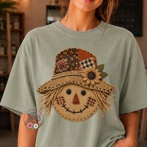 May include: A sage green t-shirt featuring a central graphic of a scarecrow face wearing a patchwork hat with a sunflower. The design uses textured patterns resembling burlap, plaid, and floral fabric, with black button eyes and stitched details.