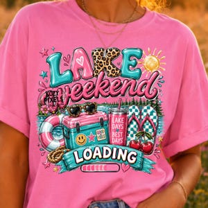 May include: A bright pink t-shirt featuring a graphic print with the text "LAKE weekend LOADING". The design includes a teal cooler, a pink and white life ring, a pink tumbler, checkered flip-flops with cherries, and a yellow sun icon.