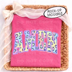 May include: A pink t-shirt with the word "AMEN" in a colorful leopard print design. The shirt is folded and presented on a woven basket, adorned with a sheer ribbon. The text "MOCK-UP INCLUDED" is also visible.