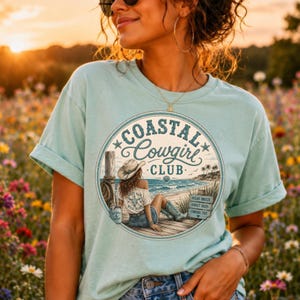 May include: A light blue t-shirt featuring a circular graphic print that displays the text Coastal Cowgirl Club. The illustration depicts a person wearing a cowboy hat and denim shorts sitting on a wooden pier overlooking the ocean.