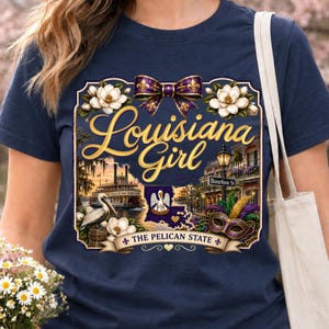 May include: A navy blue t-shirt featuring a graphic print with the text Louisiana Girl and The Pelican State. The design includes a purple and gold bow, white magnolia flowers, a steamboat, a pelican, a Bourbon St sign, and a masquerade mask.