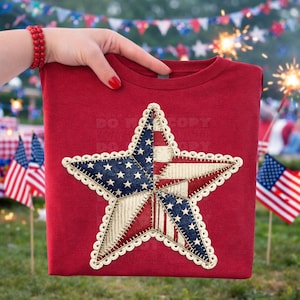 May include: A red t-shirt featuring a large star design with a patriotic theme. The star is divided into sections with the American flag's colors and patterns, including blue with white stars, red stripes, and white sections. The star is outlined with a decorative trim.