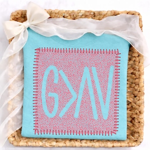 May include: A light blue t-shirt with the text "G>N" in a light blue color, stitched onto a pink patterned background. The shirt is presented in a woven basket, adorned with a white ribbon.