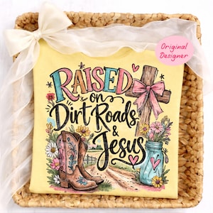 K&ouml;nnte beinhalten: Ein hellgelbes T-Shirt mit einem Grafikdruck und dem Text "Raised on Dirt Roads & Jesus". Das Design zeigt ein braunes Holzkreuz mit einer rosa Schleife, ein Paar braune Cowboystiefel, ein blaues Einmachglas mit Blumen und kleine Herzen.