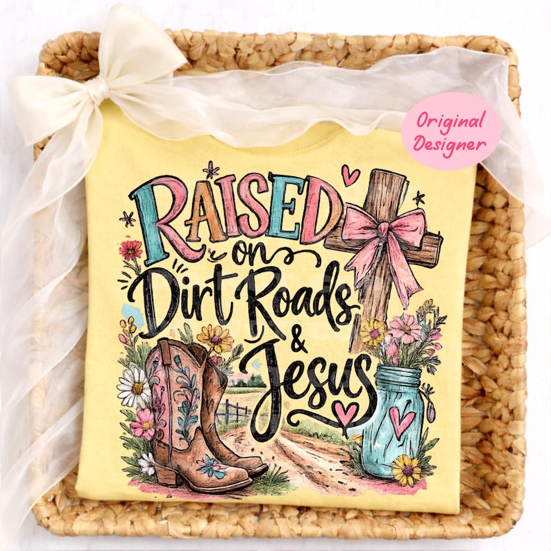 K&ouml;nnte beinhalten: Ein hellgelbes T-Shirt mit einem Grafikdruck und dem Text "Raised on Dirt Roads & Jesus". Das Design zeigt ein braunes Holzkreuz mit einer rosa Schleife, ein Paar braune Cowboystiefel, ein blaues Einmachglas mit Blumen und kleine Herzen.