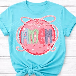 May include: A light blue t-shirt featuring the word "AMEN" in colorful letters within a pink, scalloped design. The design includes a pink bow and small flowers. The shirt is tied at the waist.