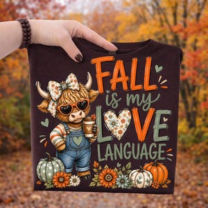 May include: A dark brown t-shirt featuring a graphic of a Highland cow wearing a floral bow, heart-shaped sunglasses, and denim overalls while holding a coffee cup. The text reads FALL is my LOVE LANGUAGE, accompanied by pumpkins and flowers.