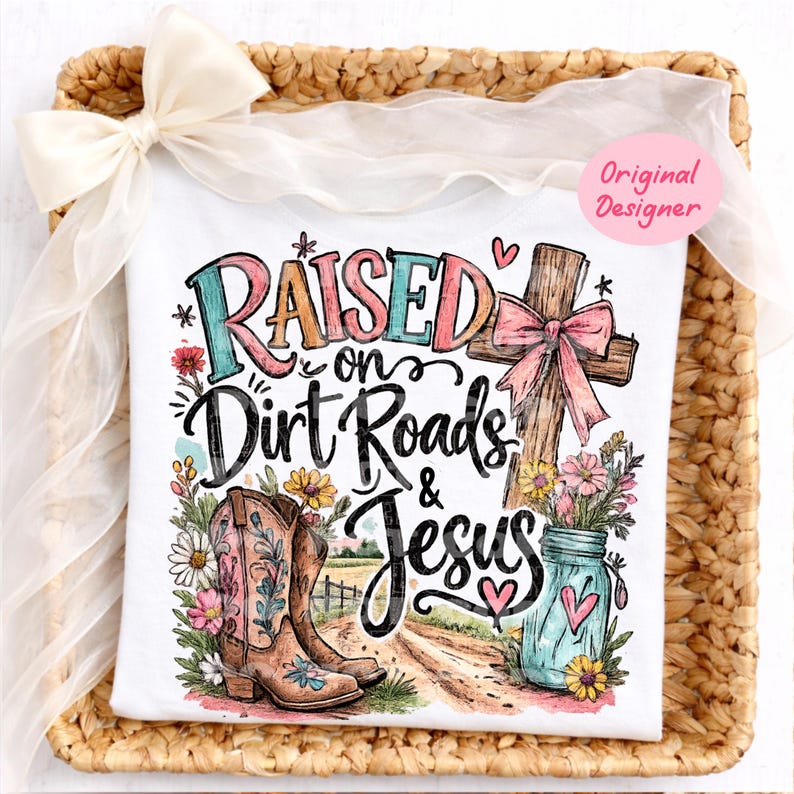 K&ouml;nnte beinhalten: Ein wei&szlig;es T-Shirt mit einem Grafikdruck und dem Text Raised on Dirt Roads & Jesus. Das Design zeigt ein Holzkreuz mit einer rosa Schleife, braune Cowboystiefel, ein blaues Einmachglas mit Blumen und eine Feldwegszene mit bunten Wildblumen.
