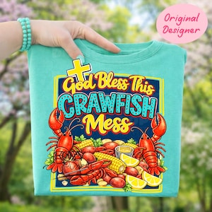 May include: A turquoise t-shirt with a colorful graphic that reads "God Bless This Crawfish Mess." The design features red crawfish, corn, potatoes, and lemons. A yellow cross is above the text. The words "Original Designer" are in a pink circle.