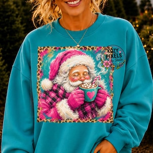 May include: A teal crewneck sweatshirt featuring a square graphic of Santa Claus wearing a pink and black plaid coat and hat, holding a teal mug with a pink heart design filled with marshmallows, surrounded by a leopard print border.