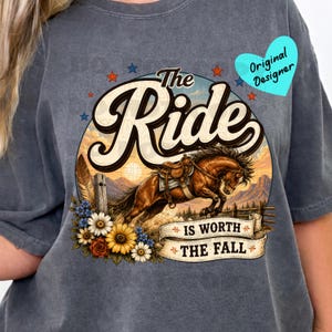 May include: Dark grey t-shirt with a graphic of a horse jumping over a fence, with the words "The Ride is Worth The Fall". The design includes a sunset, flowers, and stars.