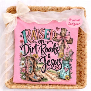 K&ouml;nnte beinhalten: Ein hellrosa T-Shirt mit einem Grafikdruck und dem Text "Raised on Dirt Roads & Jesus". Das Design zeigt ein Holzkreuz mit einer rosa Schleife, ein Paar braune Cowboystiefel, ein blaues Einmachglas mit Blumen und eine Feldweg-Szene.