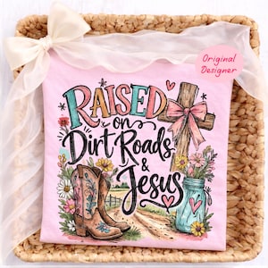 K&ouml;nnte beinhalten: Ein hellrosa T-Shirt mit einem Grafikdruck und dem Text "Raised on Dirt Roads & Jesus". Das Design zeigt ein Holzkreuz mit einer rosa Schleife, braune Cowboystiefel, ein blaues Einmachglas mit Blumen und eine Feldweg-Landschaft.
