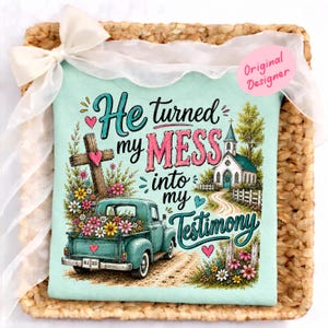 Könnte beinhalten: Hellblaues T-Shirt mit dem Text "He turned my MESS into my Testimony". Das Design zeigt einen türkisfarbenen Pickup-Truck voller Blumen, ein Holzkreuz und eine weiße Kirche. Das Shirt ist mit Herzen und Blumen verziert.
