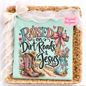 K&ouml;nnte beinhalten: Ein helles mintgr&uuml;nes T-Shirt mit einem Grafikdruck und dem Text "Raised on Dirt Roads & Jesus". Das Design zeigt ein Holzkreuz mit einer rosa Schleife, braune Cowboystiefel mit Blumenakzenten und ein blaues Einmachglas voller Blumen.