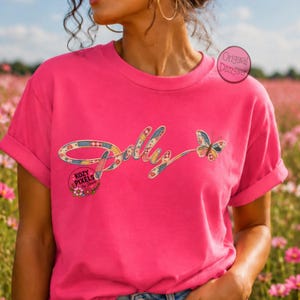 May include: A bright pink short-sleeved t-shirt featuring the word Dolly in a multicolored patchwork pattern with a matching butterfly graphic. The design includes a circular logo with the text Kozy Pixels by Sheila and an Original Designer stamp.