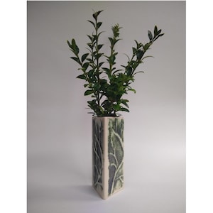 May include: A white ceramic vase with a green leaf design. The vase is filled with green leafy stems.