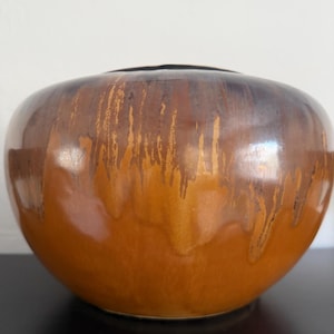 May include: A round ceramic vase with a smooth, glossy finish. The upper portion features a dark brown glaze that transitions into a lighter, warm amber-orange tone toward the base, with vertical drip patterns creating a textured visual effect.